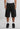 Eine Person in einem weißen Tank Top, 2Y Studios Hogan Worker Shorts Washed Black von 2Y Studios, weißen Socken und schwarzen Turnschuhen steht vor einem schlichten Hintergrund.