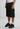 Eine Person in einem weißen Tank Top, 2Y Studios Hogan Worker Shorts Washed Black, weißen Socken und schwarzen Turnschuhen steht vor einem schlichten Hintergrund.