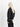Eine Person mit langem, hellblondem Haar steht seitlich und trägt eine schwarze Alpha Industries MA-1 Heritage Jacke, eine schwarze Hose und ein weißes Hemd mit asymmetrischem Saum.