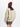 Eine Person in der beigen Bio-Jacke MA-1 Heritage von Alpha Industries, einem olivgrünen Rock und einer kastanienbraunen Mütze blickt vor einem weißen Hintergrund von der Kamera weg und verkörpert den Stil nachhaltiger Militärbekleidung.