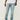 Eine Person, die True Religion Ricky SN Flap 32 IN Brilliant Medium Wash Jeans von True Religion, ein weißes T-Shirt und weiße Turnschuhe trägt, steht vor einem schlichten hellen Hintergrund.