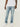 Eine Person, die True Religion Ricky SN Flap 32 IN Brilliant Medium Wash Jeans von True Religion, ein weißes T-Shirt und weiße Turnschuhe trägt, steht vor einem schlichten hellen Hintergrund.